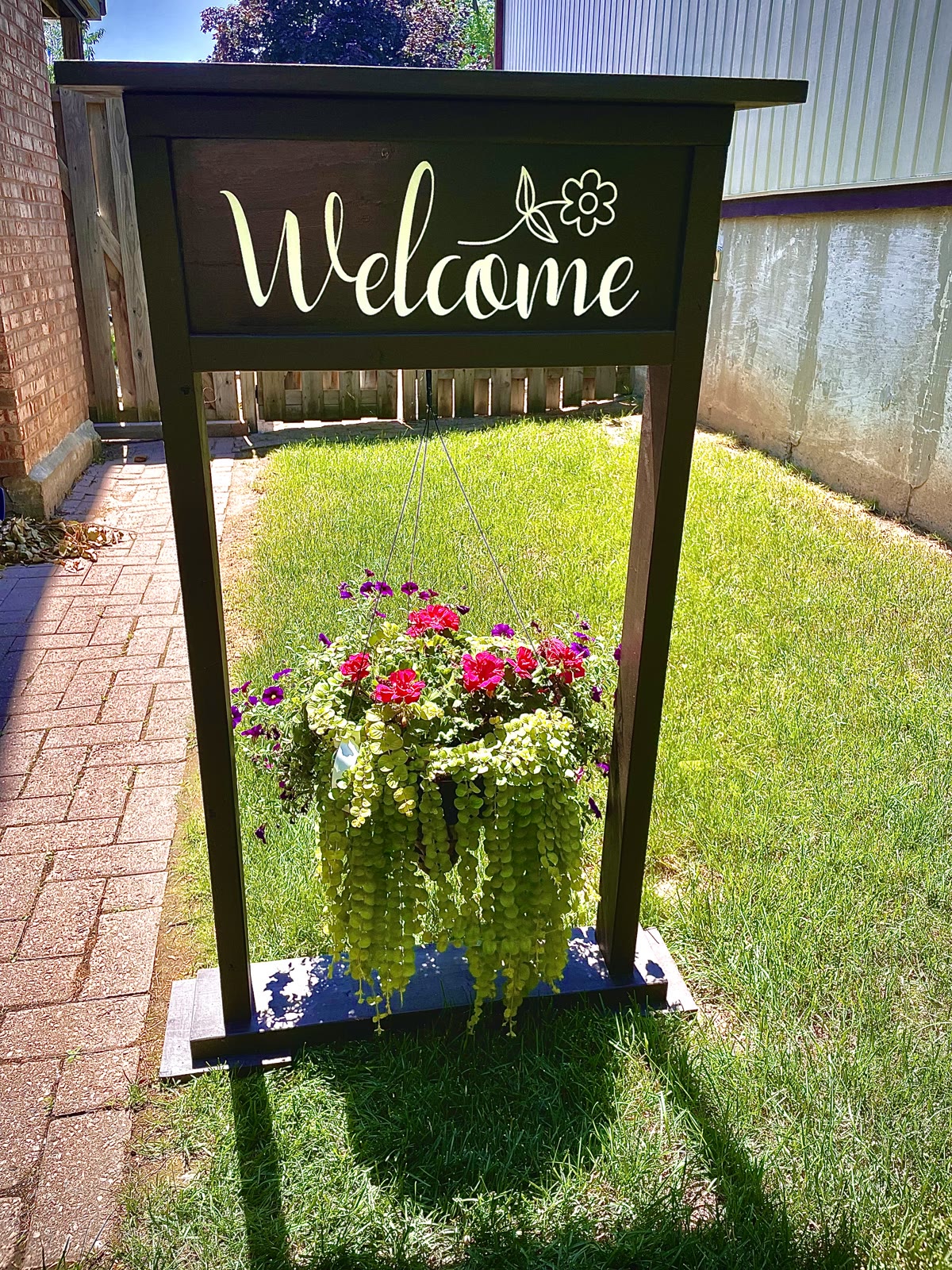 Finished black wooden welcome sign and plant holder with white vinyl lettering and a colourful hanging flower basket